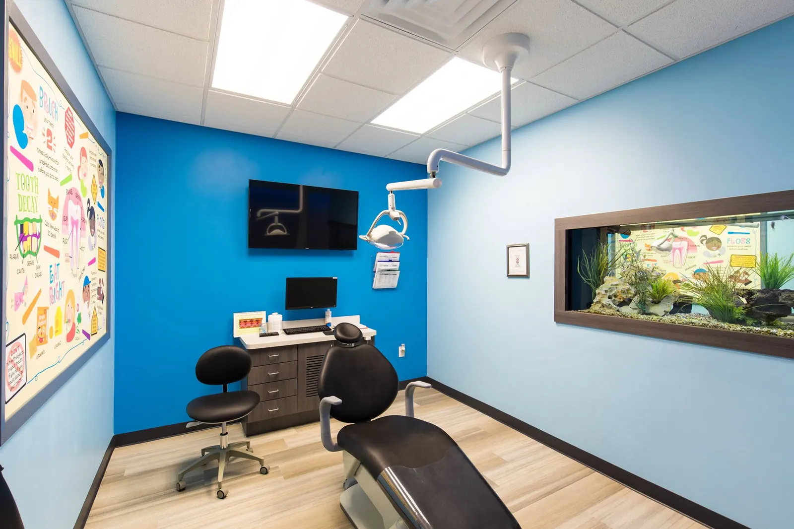 Blue treatment room with a dental chair, wall-mounted screen, and aquarium window